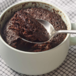 Metal spoon scooping rich, moist chocolate mug cake from a pale green ceramic mug resting on a checkered napkin